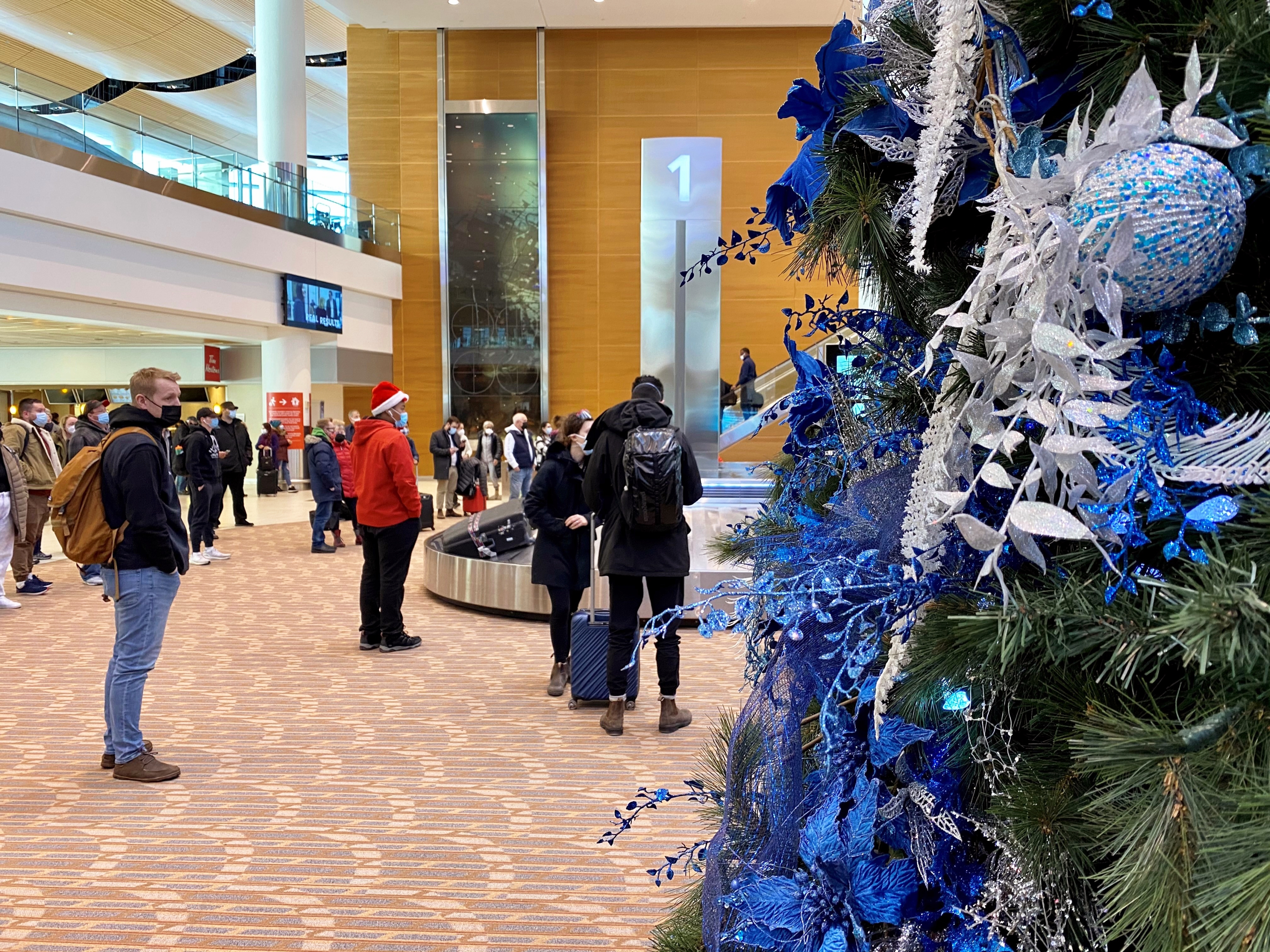 Des voyageurs se tiennent à côté d'un arbre de Noël dans le hall des arrivées en attendant leurs bagages.