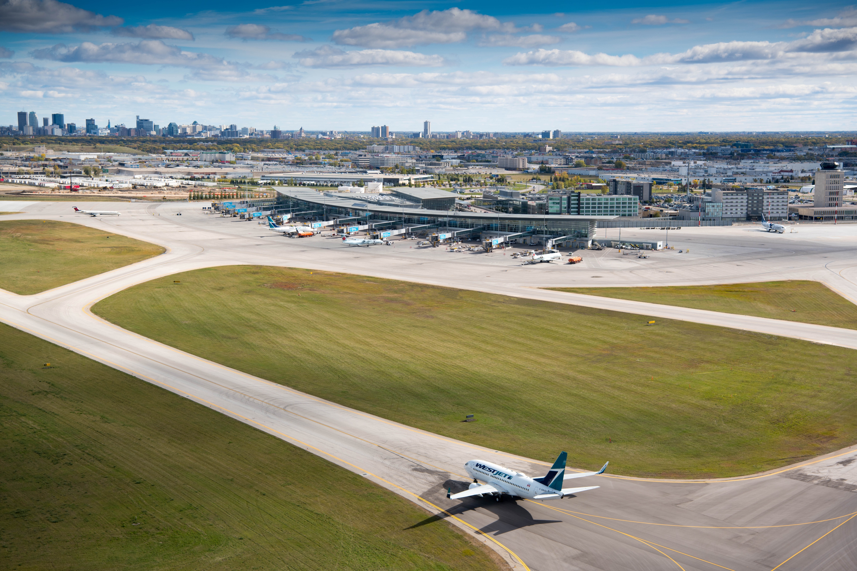 A WestJet plane taxis towards the Winnipeg Richardson International Airport terminal with the city skyline in the background.