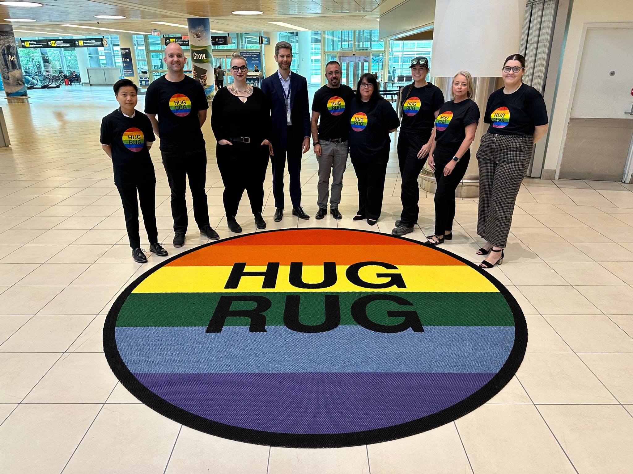 WAA employees are shown standing around a pride-themed Hug Rug in the Arrivals Hall of Winnipeg Richardson International Airport.
