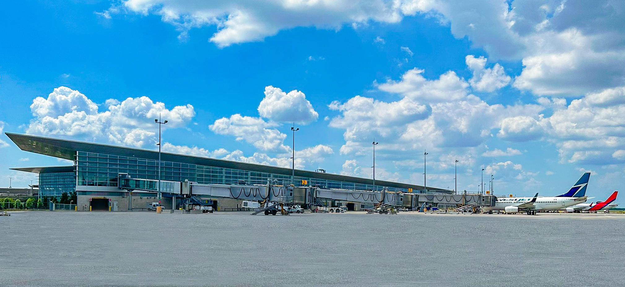 Under a blue sky with some clouds, two planes are parked at the boarding bridges of the Winnipeg Richardson International Airport terminal with the main apron shown in the foreground.