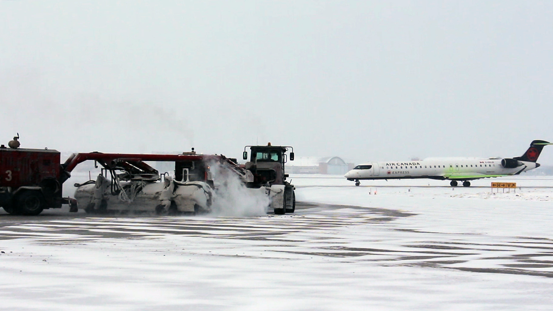 A plow clears snow on the airfield as an Air Canada plane taxis in the background.