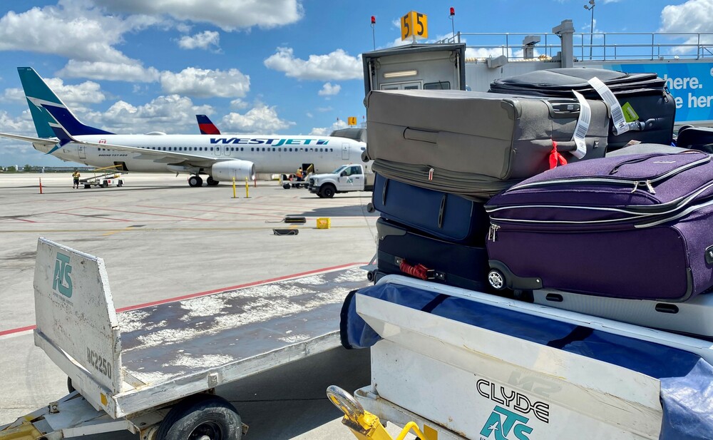 Bags are stacked on a luggage cart, waiting to be loaded on a plane with a WestJet aircraft in the background.